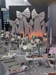 Mewujudkan acara impian Anda bersama Vendor Dekorasi profesional yang siap menghadirkan estetika terbaik di setiap sudut ruangan. Elegant banquet table set with pastel pink and white flowers, tall candles, and a backdrop of large gray bows for a formal event.